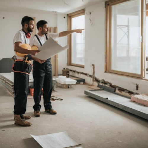 Full length of construction workers analyzing blueprints in the apartment while one of them is aiming at distance.